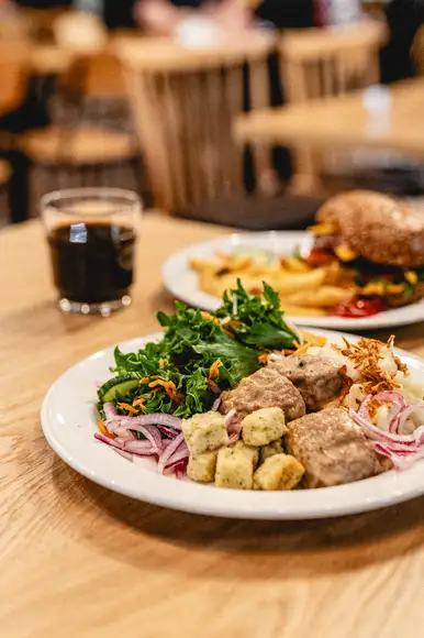 Lähikuva lounasannoksesta, jossa Ståhlbergin omia lihapullia kermakastikkeessa, krutonkeja, slaattia ja perunamuusia. Taustalla lasi kotikaljaa ja lautanen jolla hampurilainen ja ranskalaisia burgerbuffasta.